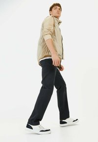 Beige lichtgewicht jack met een rits aan de voorkant, gecombineerd met donkerblauwe broek en witte sneakers met marineblauwe accenten, staande tegen een witte achtergrond.