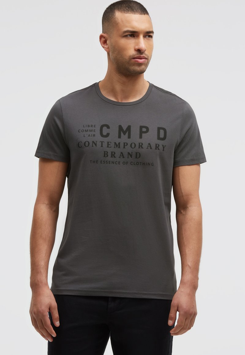 Man with short hair wearing dark gray T-shirt with black “CMPD Contemporary Brand” text, standing against a plain light background.