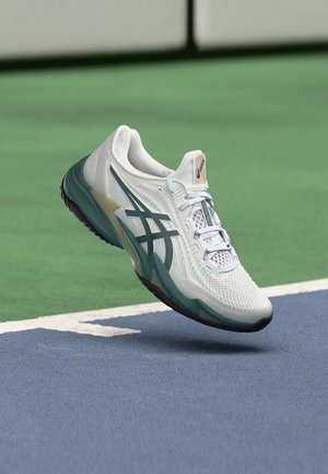 Witte en groene ASICS hardloopschoen die boven een blauw-groene tennisbaan zweeft vlakbij de lijn.