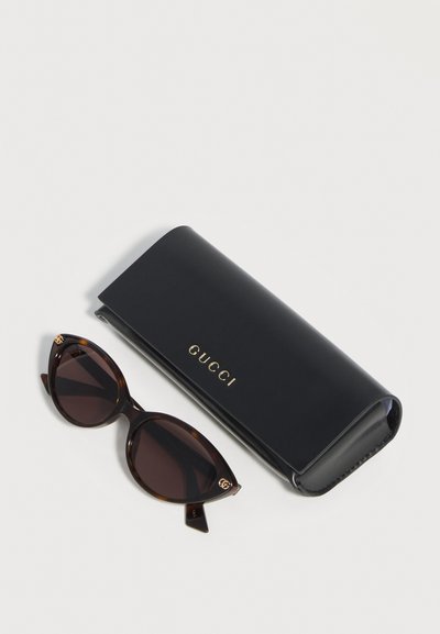 Γυαλιά ηλίου τύπου cat-eye σε ταρταρούγα με σκοτεινούς φακούς δίπλα σε μαύρη θήκη Gucci με χρυσό λογότυπο σε λευκό φόντο.