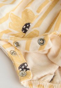 Žltá a krémová prúžkovaná látka s kvetinovými vzormi, zdobená čierno-bielymi lienkami a striebornými patentkami. Mäkká textúra.