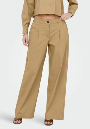 Beżowe, szerokie spodnie z wysokim stanem i plisami z przodu, zapinane na guziki, noszone z dopasowaną, krótką koszulą z długim rękawem oraz brązowymi sandałami z odkrytymi palcami.