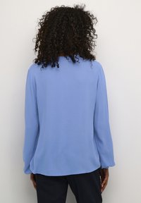 Haut à manches longues en bleu clair, en tissu texturé avec une coupe décontractée. Il présente un col rond et des manches légèrement évasées.