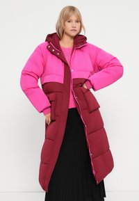 Duga puffer jakna s ružičastim gornjim dijelom i burgundskim donjim dijelom, ima kapuljaču, kopčanje na drukere, dva prednja džepa i rebraste manžete.