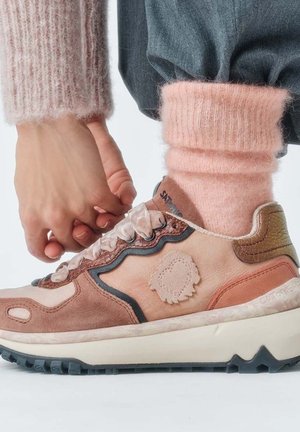 Persona con calzini rosa pelosi e sneakers beige e marroni che si china per regolare i lacci della scarpa del piede destro.