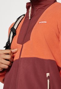 Giacca in pile con blocchi di colore arancione e bordeaux, colletto alto, chiusura a mezza zip e dettaglio del logo sul petto.