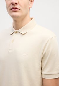 Beige színű, pamut polo ing klasszikus gallérral, két gombbal és diszkrét, bélyegzett "BOSS" logóval a mellkason. Sima textúra.