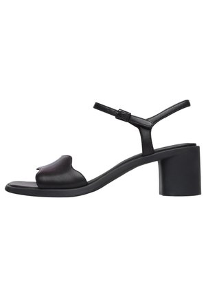 Sandalias de tacón de cuero negro con una parte frontal curva, correa ajustable en el tobillo y tacón grueso. Textura suave y diseño minimalista.