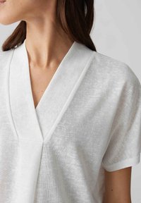 Top bianco a maniche corte in tessuto leggero con scollatura a V e una sottile texture. Presenta linee pulite e una vestibilità rilassata.