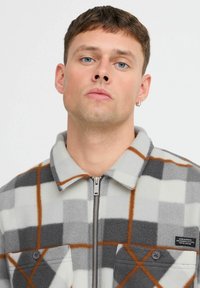 Chaqueta de forro polar a cuadros en gris y blanco con detalles en naranja, cremallera y dos bolsillos delanteros. Textura suave y diseño con cuello.