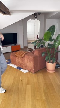 Kollane korduroy diivan ümarate vormidega, pruun viskamine ja suur roheline taim terrakotapottides. Puitpõrand kõikjal.