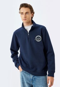 Sudadera de felpa azul marino con cremallera de 1/4 y textura suave; presenta un gráfico de "Aventura" en el lado izquierdo del pecho y puños acanalados.