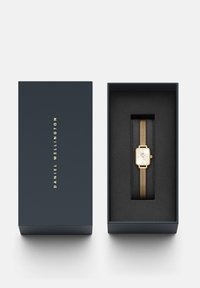 Goldmesh-Armbanduhr mit rechteckigem weißen Ziffernblatt in einer marineblauen Box. Das Innere der Box ist aus schwarzem Samt; der Markenname ist in Gold auf der Außenseite der Box geprägt.