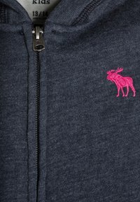 Donkerblauwe ritssluiting kindervest met een klein roze geborduurd elkemotief op de borst en maatlabel zichtbaar binnenin de kraag.