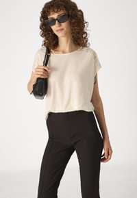 Beige top s krátkým rukávem a krajkovými akcenty na ramenou, spárovaný s černými kalhotami s vertikálními švy, a drží malou černou tašku.