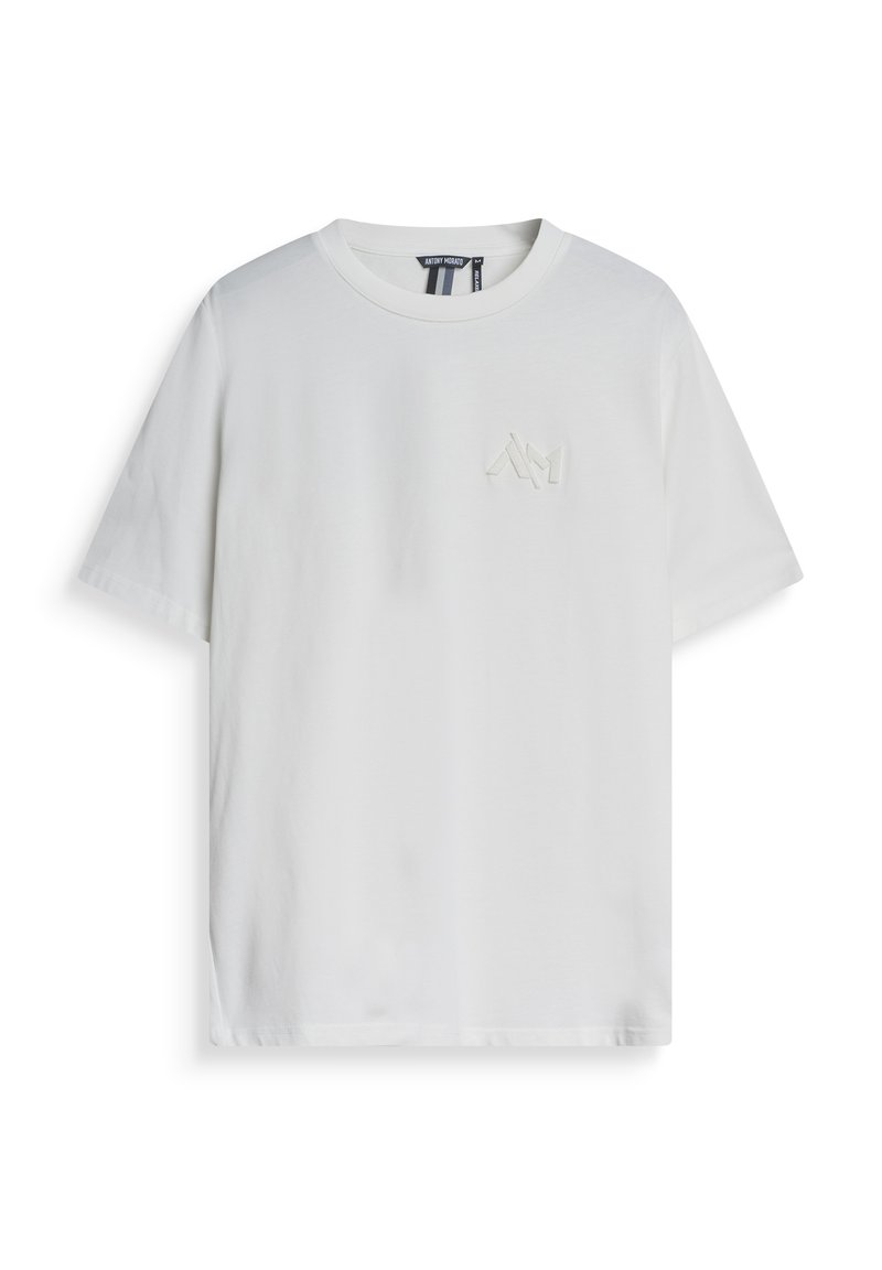 Camiseta de algodón blanco con mangas cortas, cuello redondo y un pequeño logo tono sobre tono en el lado izquierdo del pecho. Textura suave, corte casual.