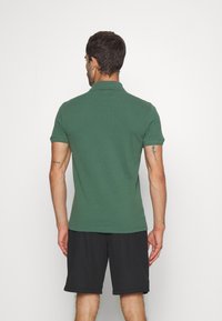 Man standing facing away wearing a green short-sleeve polo shirt and black shorts, showing tattoo details on his arms.