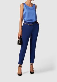 Femme portant un haut bleu en satin sans manches, un pantalon bleu taille haute avec une ceinture chaîne, tenant une pochette noire et portant des escarpins noirs à bride autour de la cheville.
