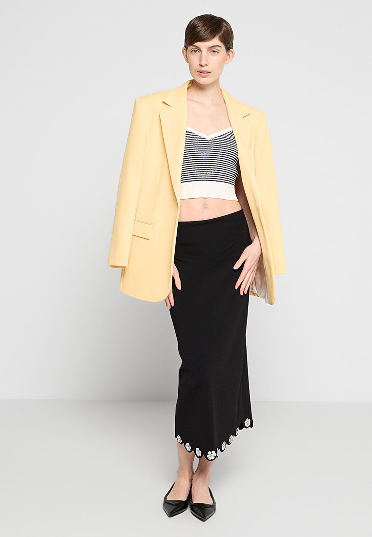 Gelber übergroßer Blazer über einem gestreiften, kurzes Oberteil und einem schwarzen, langen Rock mit floralen Stickereien. Dazu elegante, spitze schwarze Schuhe.
