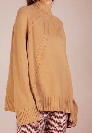 Femme portant un pull à col roulé ample de couleur camel avec poignets et ourlet côtelés, associé à un pantalon à carreaux rouge, blanc et bleu.