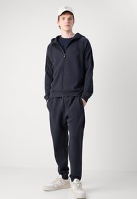 Mornarska zip-up dukserica s podesivom špagom, uparena s odgovarajućim trenirkama. Sadrži logo na prsima i bočne džepove, nosi se s bijelim tenisicama.