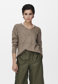 Brauner gerippter Strickpullover mit V-Ausschnitt, langen Ärmeln und leicht übergroßer Passform, kombiniert mit hochgeschnittenen grünen Hosen.