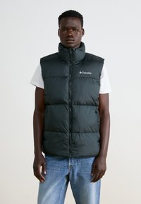 Sort puffy vest med høj krave, frontlynlås og Columbia-logo; båret over en hvid t-shirt. Blå jeans fuldender outfittet.