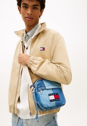 Jaunas vyras dėvi smėlio spalvos striukę ir dryžuotą marškinius, nešiojasi mėlyną derinio stiliaus džinsinį peties krepšį su Tommy Jeans logotipu.