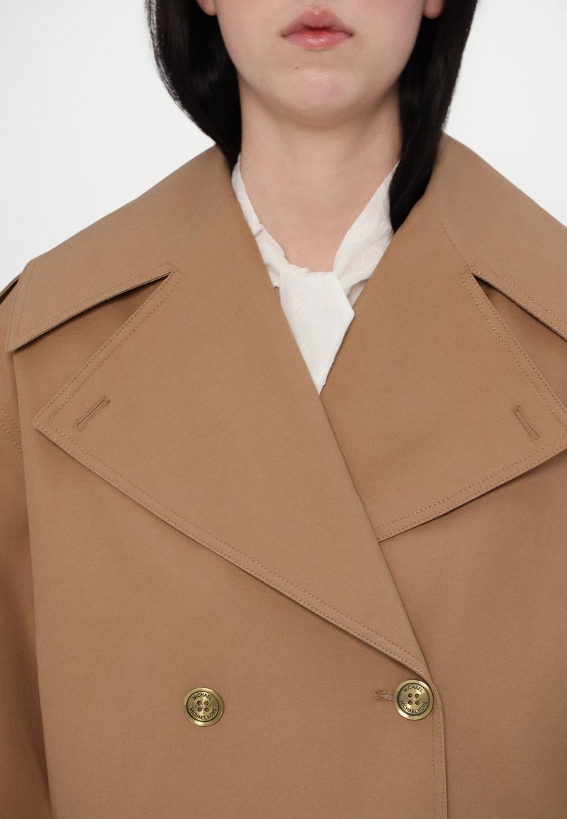 Beżowy płaszcz dwurzędowy z dużymi klapami i złotymi guzikami, prezentujący dopasowany krój i gładką teksturę tkaniny.