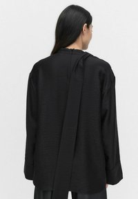 Sort langærmet bluse med en struktureret overflade, der har et bånd i ryggen og en afslappet pasform, designet med en rund udskæring og brede manchetter.