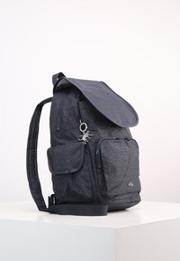 Czarny plecak z materiału o teksturowanym wzorze, z klapą zamykającą, boczną kieszenią i ozdobnym brelokiem. Regulowany pasek dla wygody.