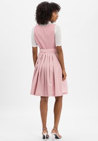 Eine Frau mit lockigem Haar trägt ein ärmelloses, rosa plissiertes Kleid über einer weißen gemusterten Bluse und silbernen High-Heels, während sie sich abwendet.