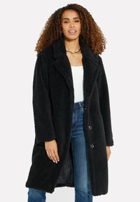 Cappotto lungo, nero e morbido con colletto, due bottoni e tasche laterali visibili, abbinato a una maglietta bianca e jeans blu.