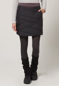 Černá nadýchaná sukně s šikmým prošíváním, elastickým pasem a bočními kapsami; kombinována se šedými punčochami a černými návleky na nohy přes boty.