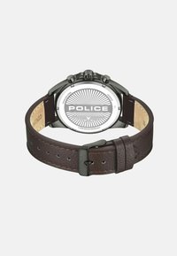 Brūns ādas siksniņas pulkstenis ar nerūsējošā tērauda korpusu, melnām akcentiem un apļveida balto ciparnīcu, kas izceļas ar saulītes rakstu un "POLICE" logotipu.