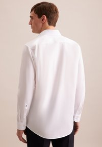 Camicia bianca a maniche lunghe in tessuto liscio, con collo classico e orlo curvo. Presenta sottili dettagli in bottoni sui polsini.