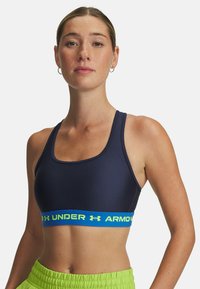 Marinblå sport-bh i mjukt tyg med ett blått elastiskt band som har "Under Armour"-text i neongrön; rundad halsringning och breda axelband.