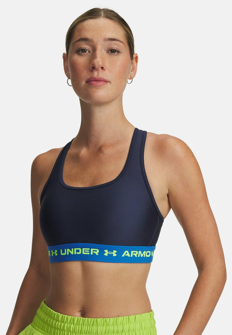 Marinblå sport-bh i mjukt tyg med ett blått elastiskt band som har "Under Armour"-text i neongrön; rundad halsringning och breda axelband.