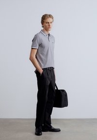 Γκρι πουκάμισο πόλο με λευκή περίμετρο, μαύρο παντελόνι και μαύρα παπούτσια. Κρατά μια μαύρη τσάντα. Απλός σχεδιασμός, μεσαία εφαρμογή, λεία υφή.