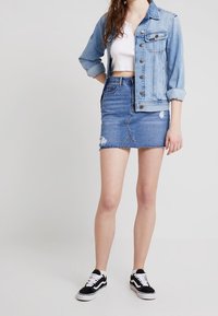 Veste en denim bleu clair, avec poches avant et fermeture à boutons. Top court blanc en dessous. Jupe en denim bleu, déchirée avec un ourlet effiloché. Baskets noires.