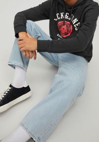 Schwarzer Hoodie mit grafischem Druck, hellblaue schmal geschnittene Jeans, schwarze Sneakers mit weißen Akzenten und weiße Knöchelsocken.