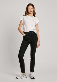 Črne jeans hlače z visokim pasom in slim fit krojem, v kombinaciji z oprijetim belim majico. Model nosi sivo športno obutev in stoji proti nevtralni ozadju.
