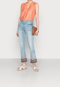 Oranssi ilman hihoja oleva paita, jossa on röyhelökaulus, vaaleansiniset farkut, joiden helmassa on brodeerattuja kuvioita, sekä beiget sandaalit, joissa on ristihihnat.