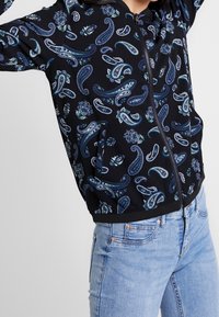 Svart zip-up jacka med paisley- och blommönster i blå och gröna toner, med huva och sidofickor, tillsammans med ljusblå jeans.