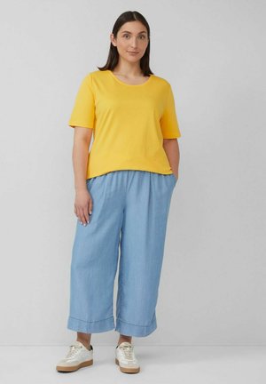 Gelbes Baumwoll-T-Shirt mit kurzen Ärmeln, kombiniert mit hellblauen weit geschnittenen Hosen, die einen Kordelzugbund und Taschen haben. Getragen mit weißen Sneakers.