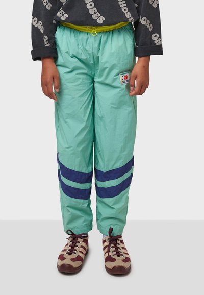 Helle tealfarbene Windbreaker-Hosen mit marineblauen Streifen. Elastischer Bund, Kordelzugverschluss und Logo-Patch. Tragen mit braunen und cremefarbenen Schuhen.