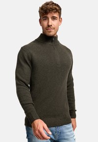 Joven vistiendo un suéter de punto con cremallera de color verde oscuro y jeans claros desgastados, de pie contra un fondo claro y liso.