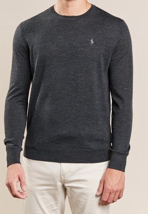 Man draagt een donkergrijze sweater met lange mouwen en een ronde hals, voorzien van een klein geborduurd logo op de borst, gecombineerd met lichtbeige broek.