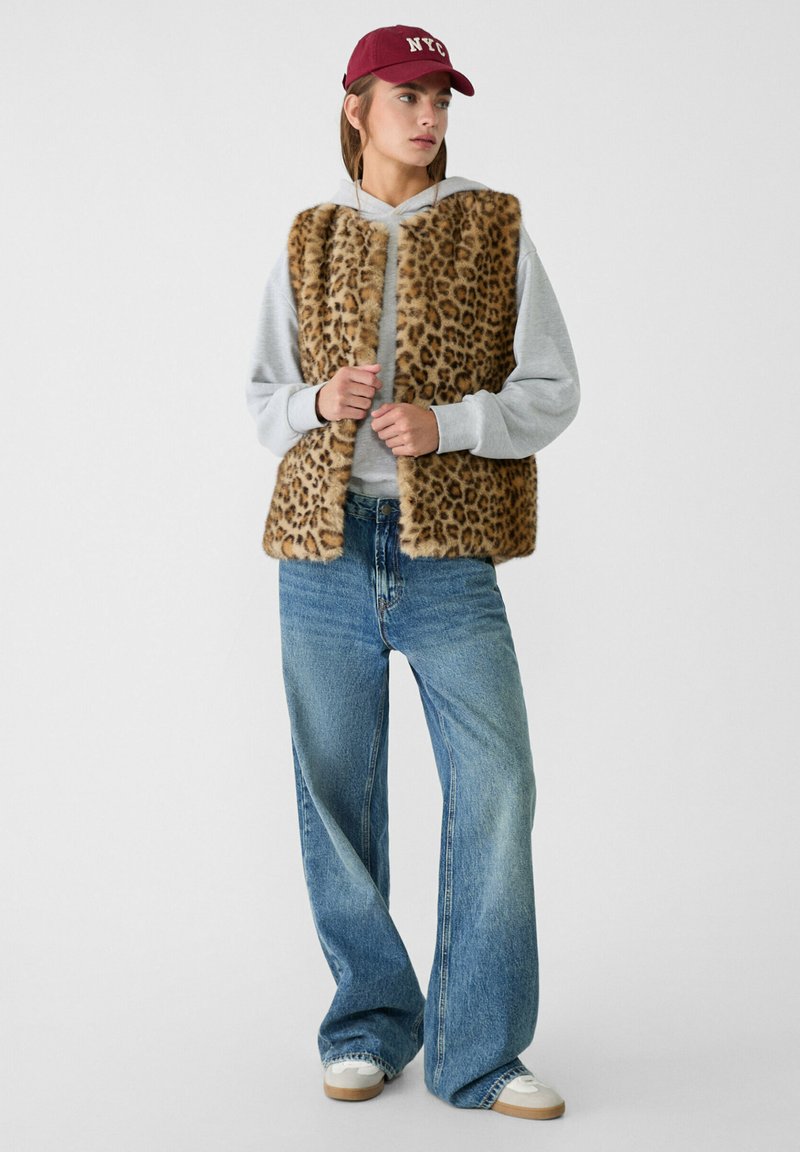 Giovane donna con cappellino rosso di New York, felpa grigia con cappuccio, gilet in pelliccia sintetica leopardata, jeans a gamba larga blu e sneakers bianche, in piedi davanti a uno sfondo bianco.