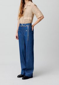 Beige-mönstrad kortärmad skjorta parat med högmidjade blå denimbyxor med vida ben som har lapplådor och ett dekorativt bälte i silverkedja.